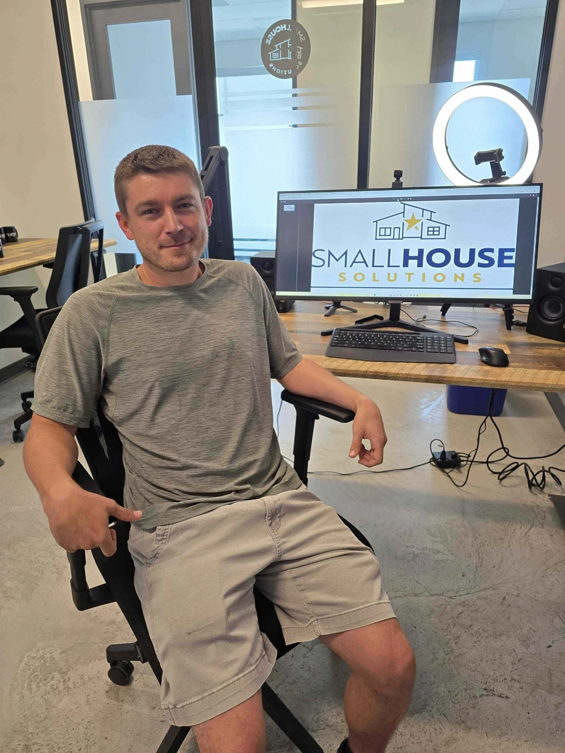 Riley Rollins, Permitting and Purchasing Coordinator at Small House Solutions, seated beside a company logo display